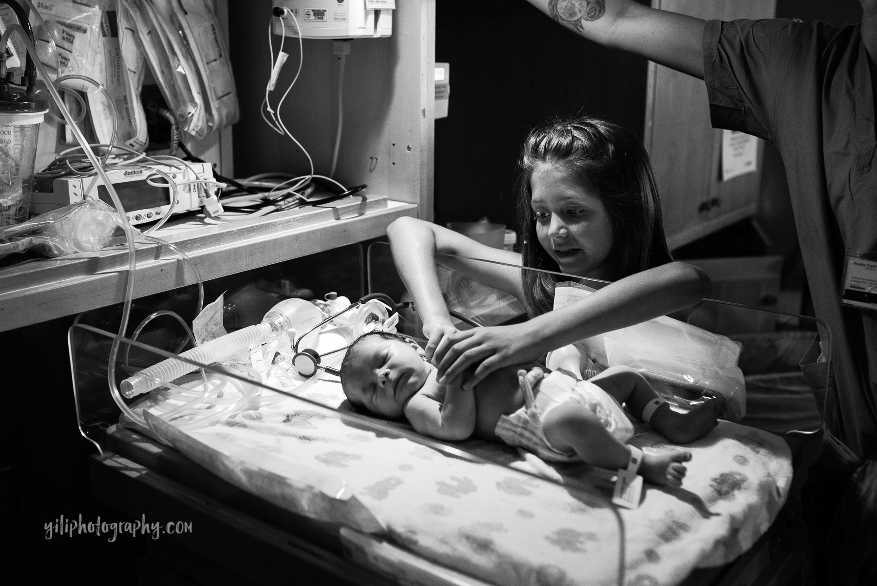 big sister holding newborn baby's arms in hospital warmer