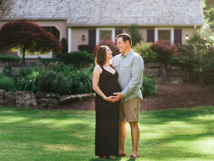 Expecting couple standing in their front yard looking affectionately at each other in golden light