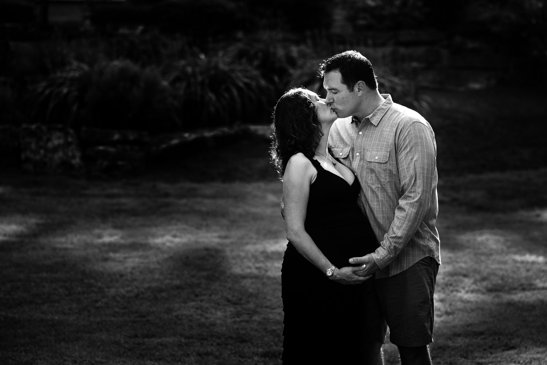 Backlit black and white of expecting couple kissing outdoors