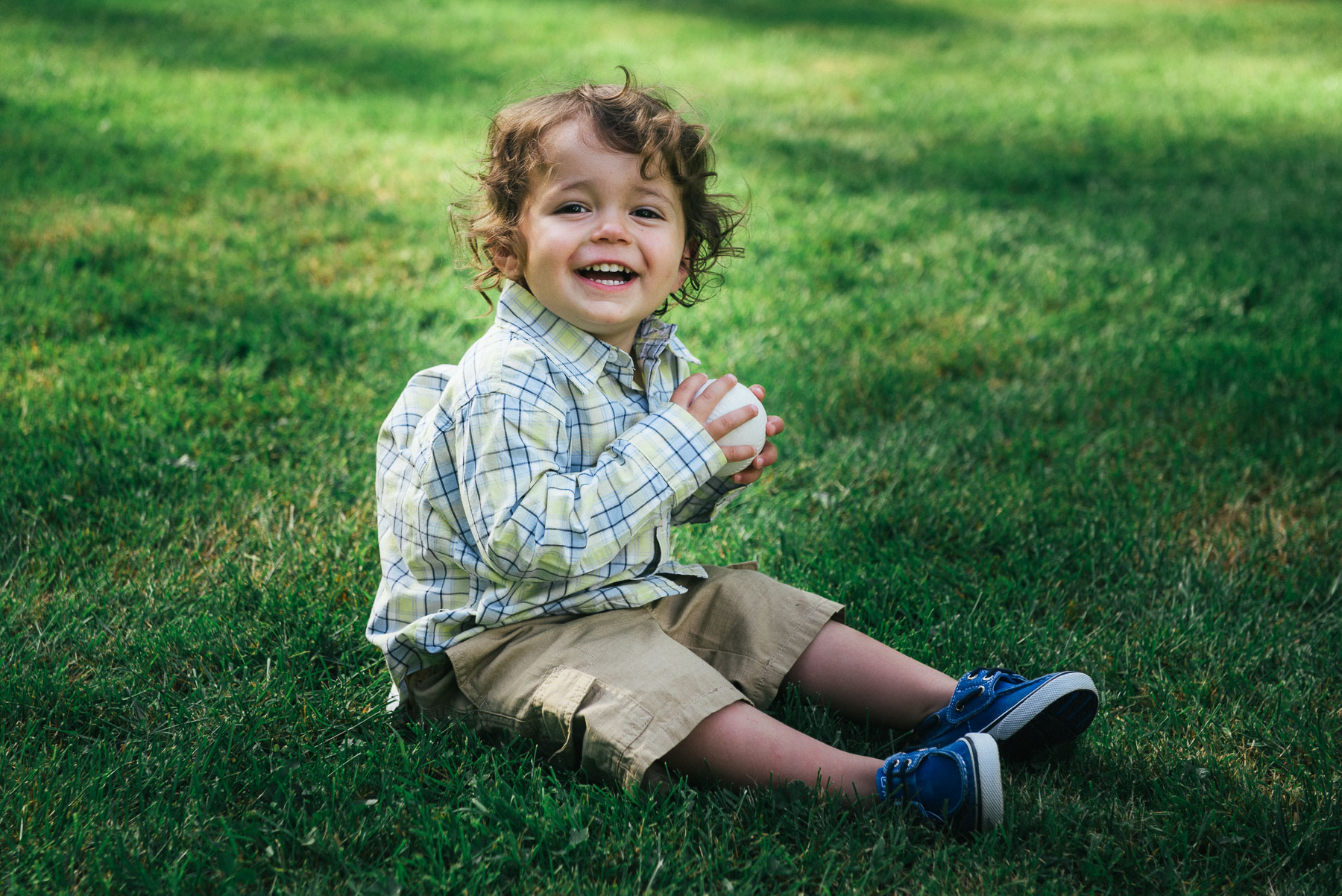 Toddler boy sitting in grass smiling and holding baseball