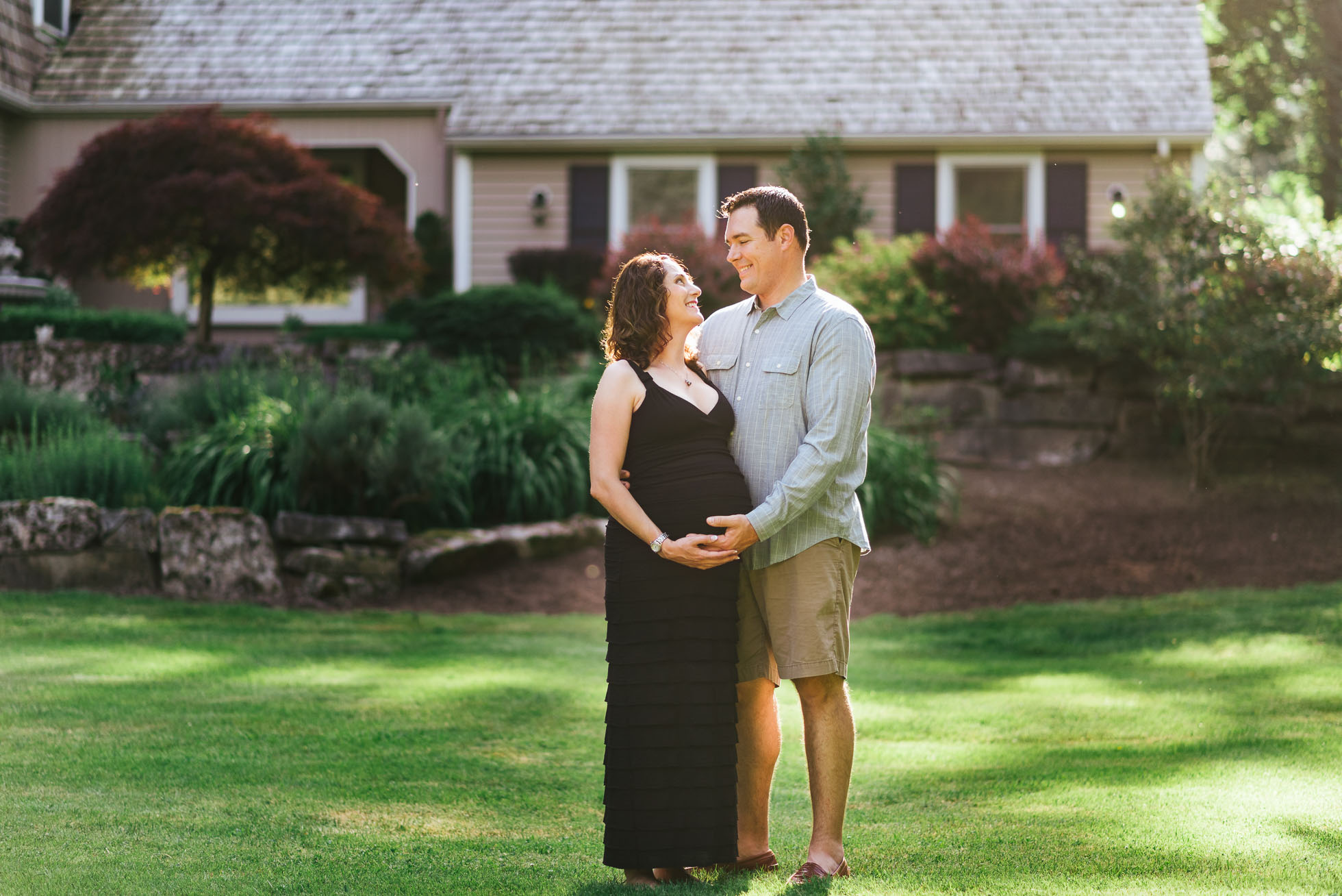 Expecting couple standing in their front yard looking affectionately at each other in golden light