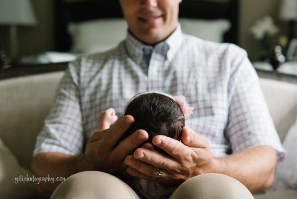 father's hands cradling newborn baby's head while seated on couch