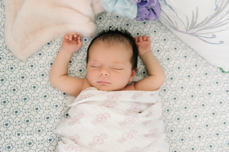 newborn brunette baby girl with arms up around head asleep in crib