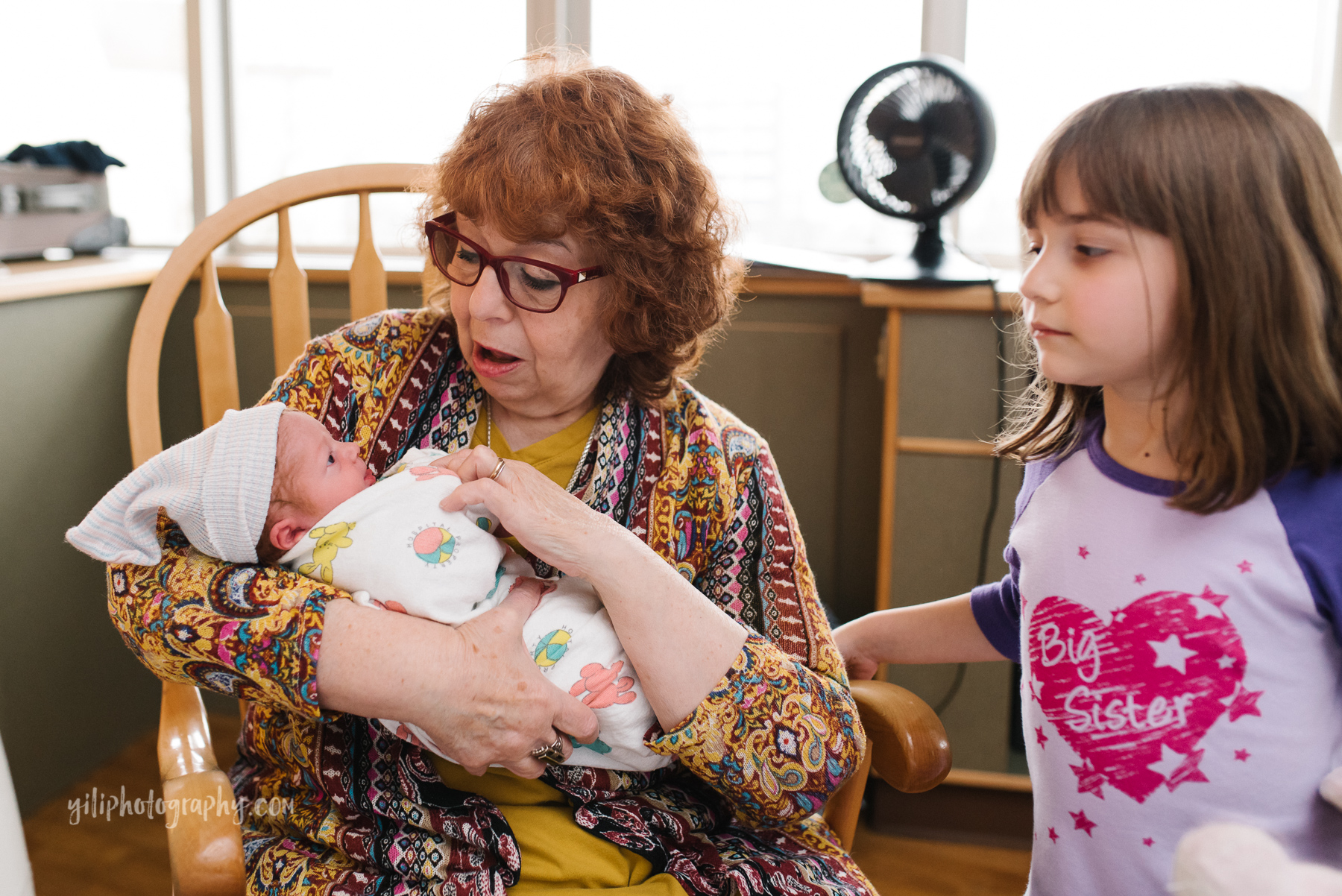 grandmother holds newborn baby while sitting in rocker in hospital room with older granddaughter