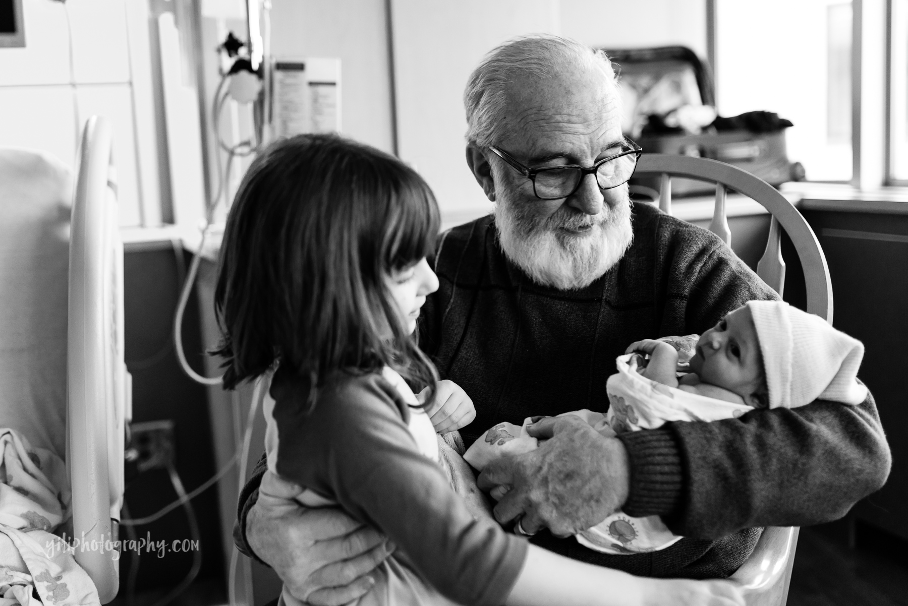 grandfather holds newborn baby in hospital rocker while hugging older granddaughter