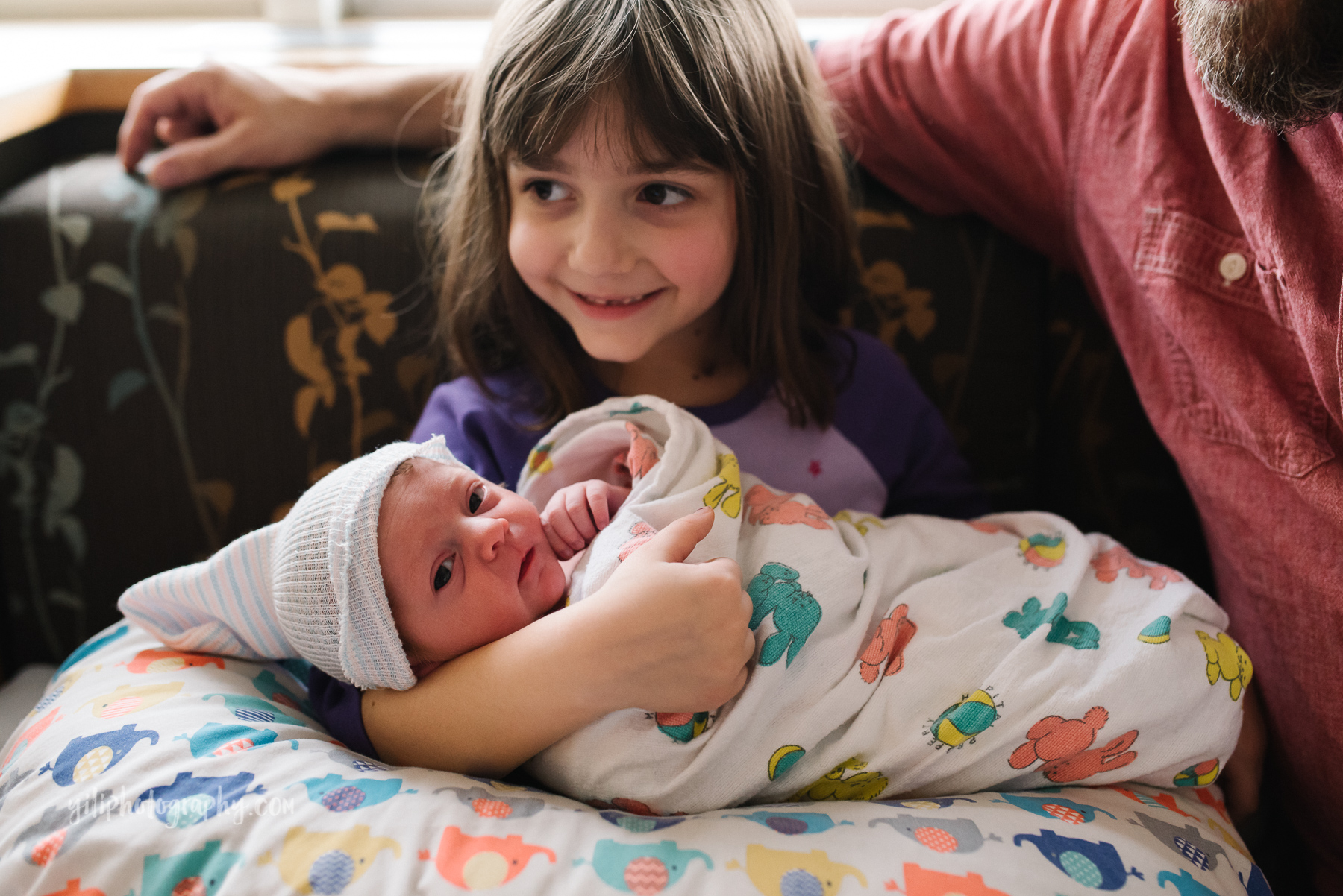 big sister sitting on hospital couch holding newborn baby sister and smiling