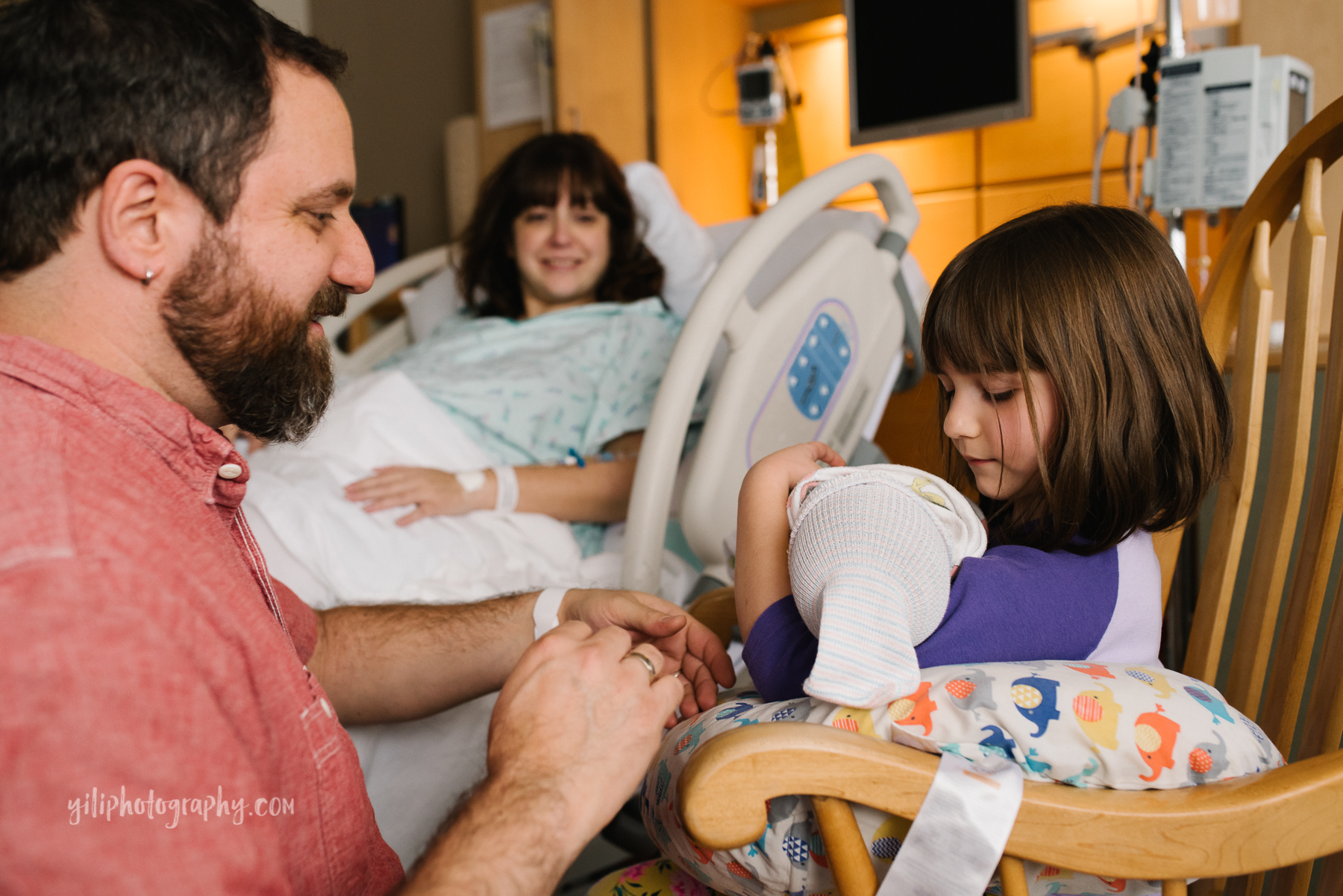 Father helps young girl hold newborn baby sister in hospital while mother looks on