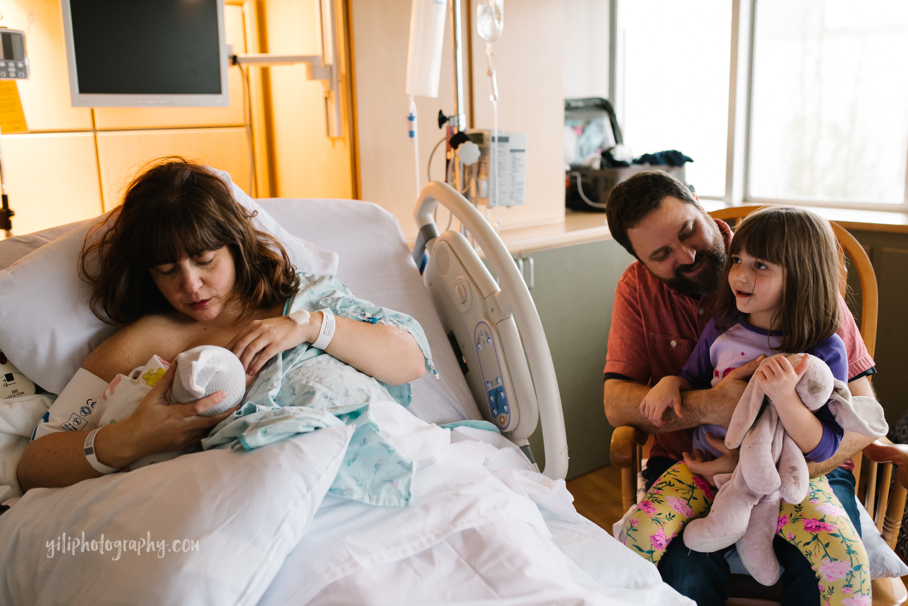 mother nurses newborn baby in hospital bed while dad and big sister look on