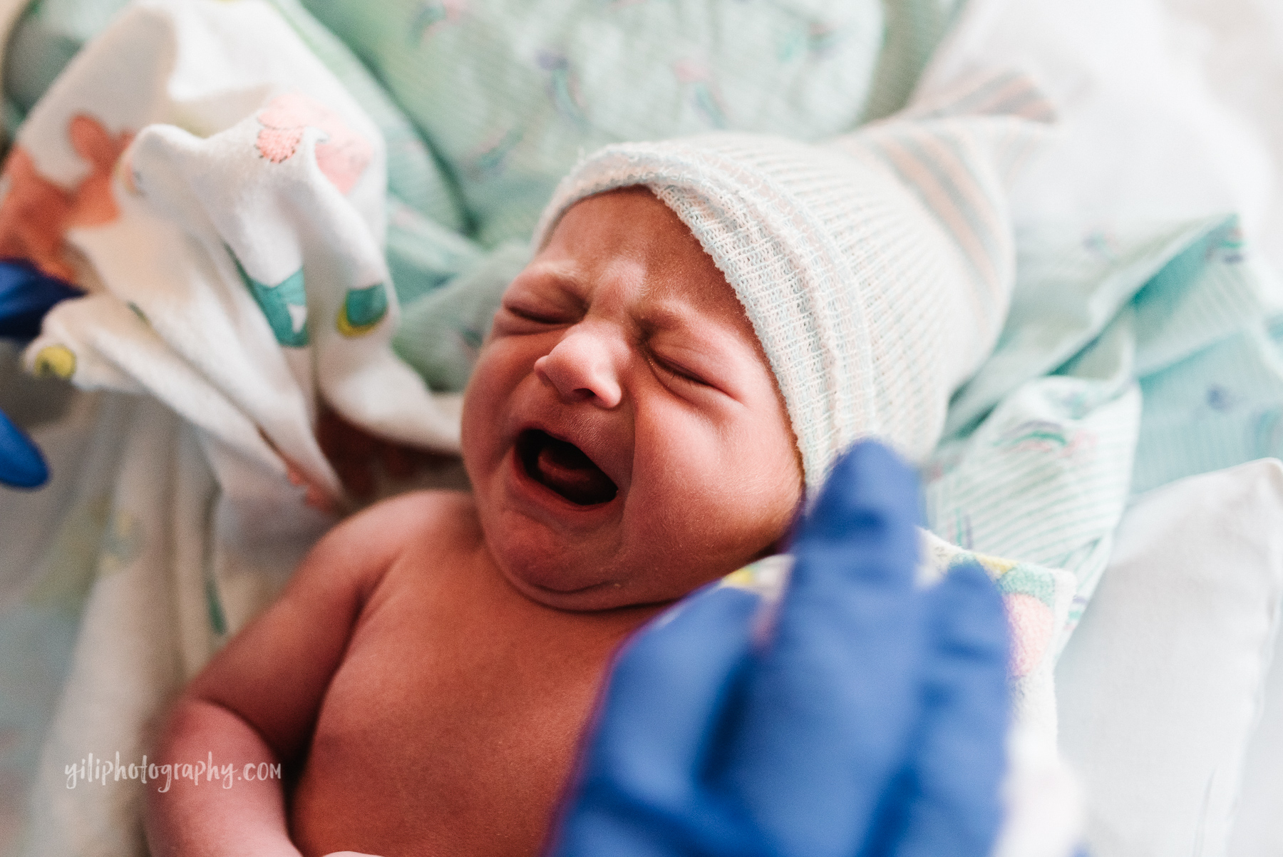 close up of newborn baby with hat crying while gloved hand assists nursing