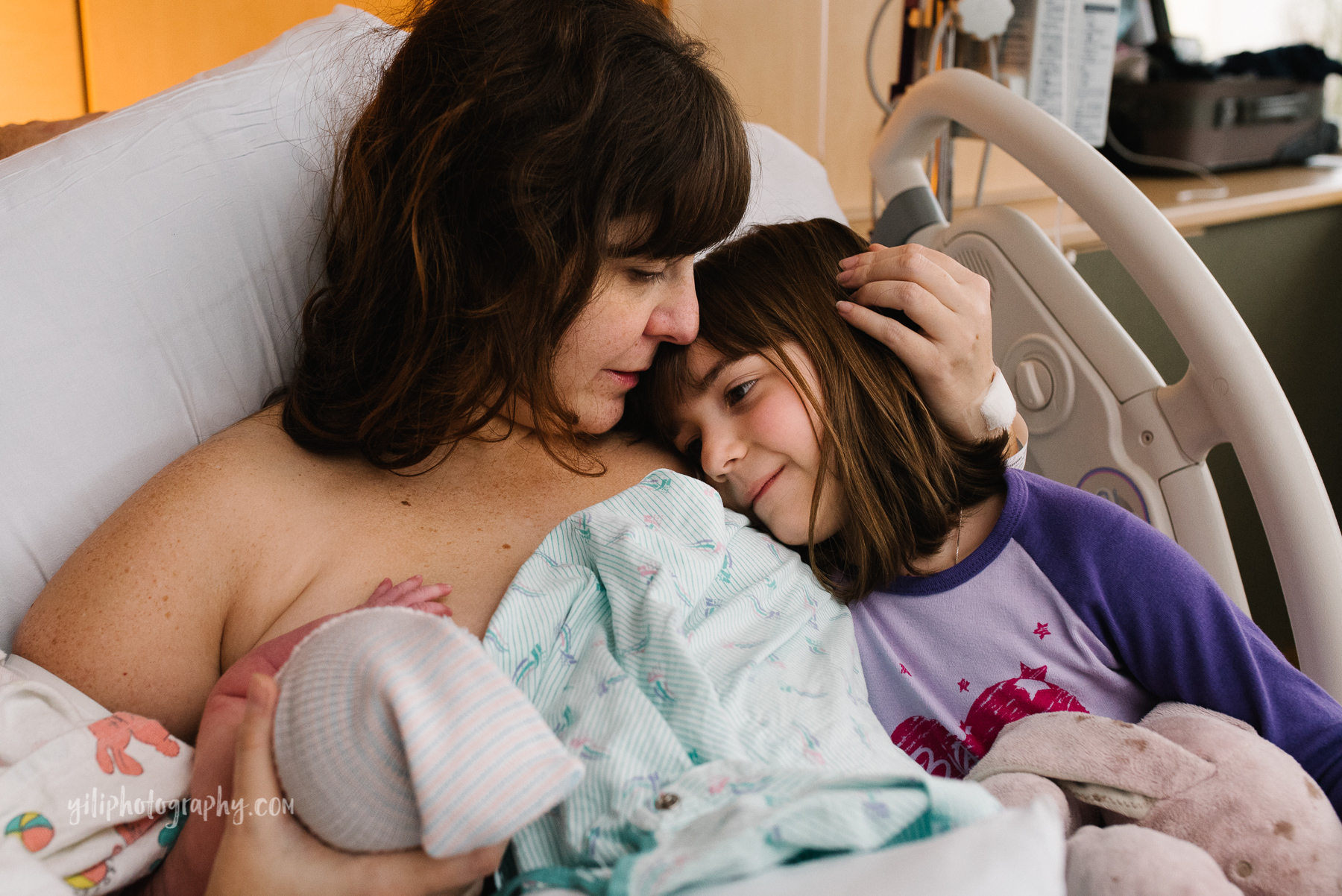 mother snuggling with her older daughter in hospital bed while nursing newborn baby