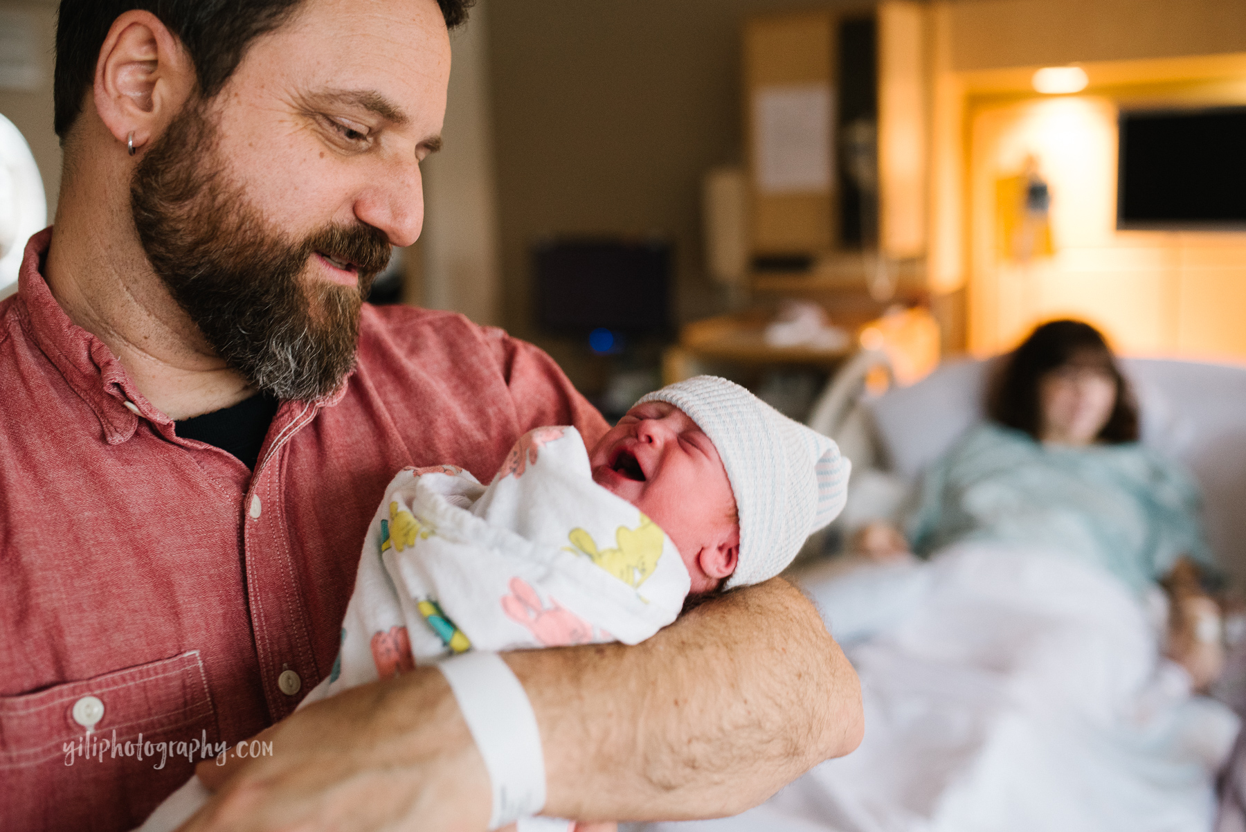 portrait of dad holding newborn baby in hospital room with mom in background