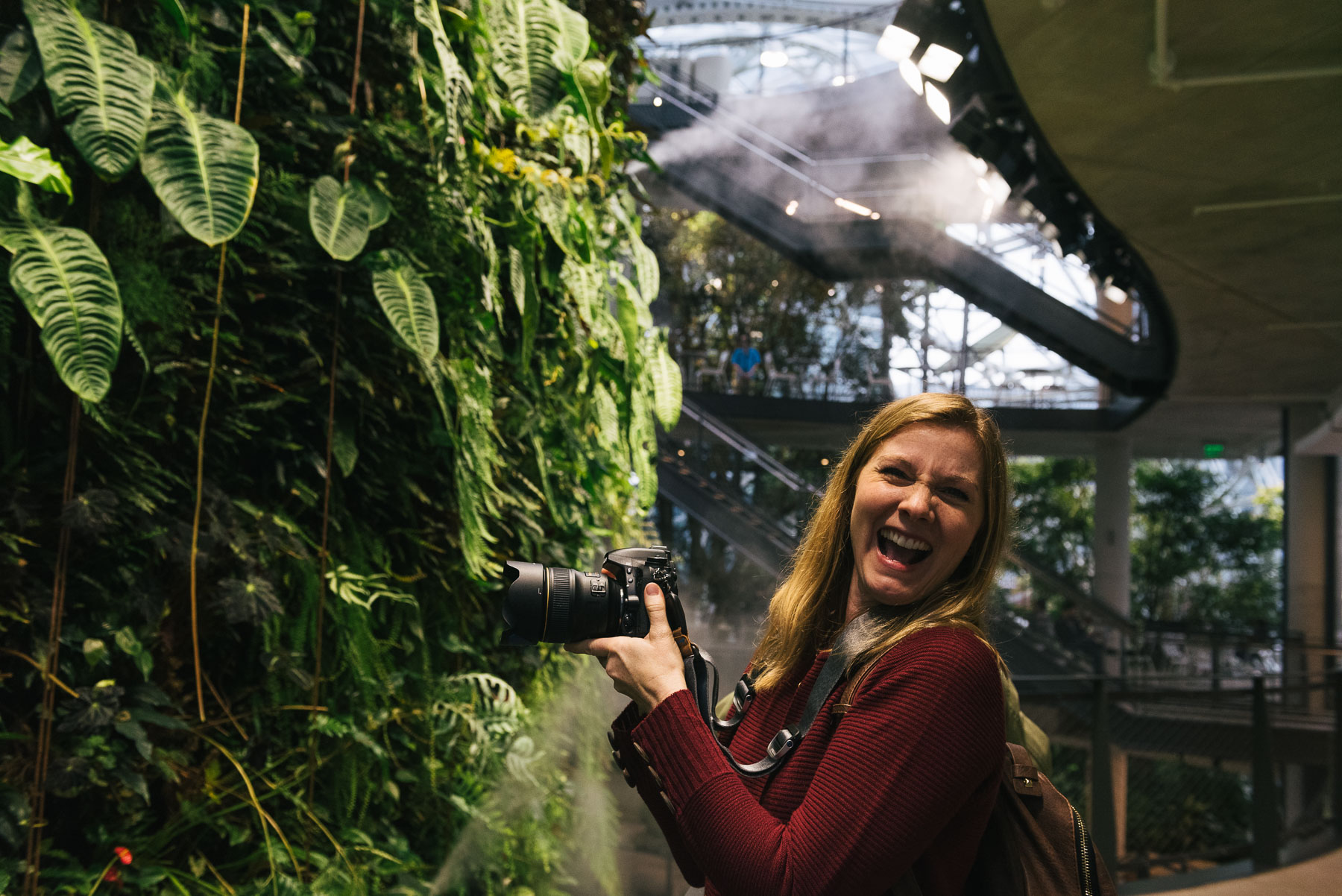 caucasian woman holding a camera and smiling while photographing plant wall inside Amazon spheres