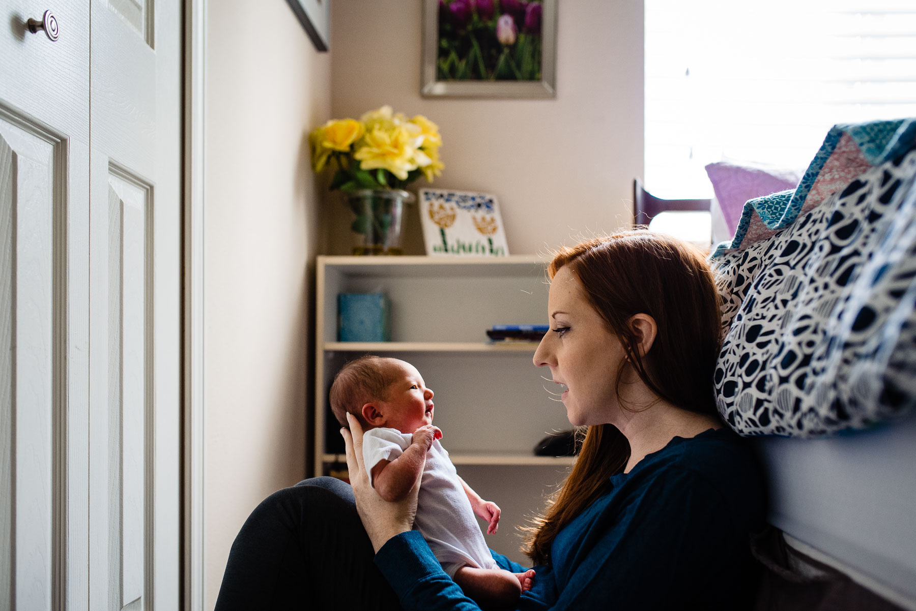 mother sitting against bed talking to newborn baby