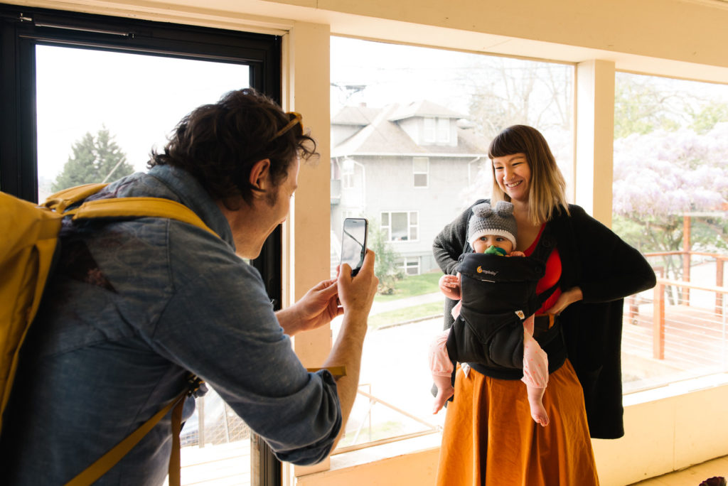 Dad photographing mom with baby in Ergo with his smart phone
